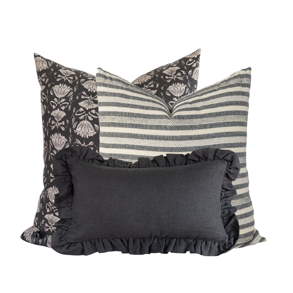 A 3 pillow combination with a square black floral pillow in the back, a square striped black pillow in the middle, and a charcoal grey lumbar pillow in from with ruffles on the perimeter.
