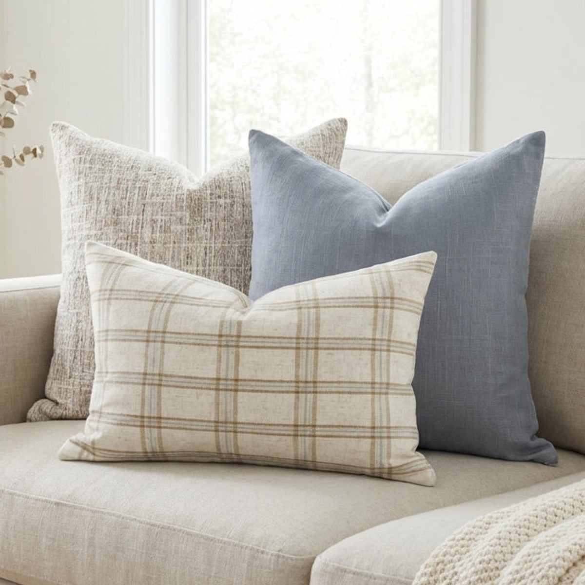 Sophisticated scene with three high-end pillow covers in a bright living room, showcasing woven texture, muted blue, and plaid pattern.