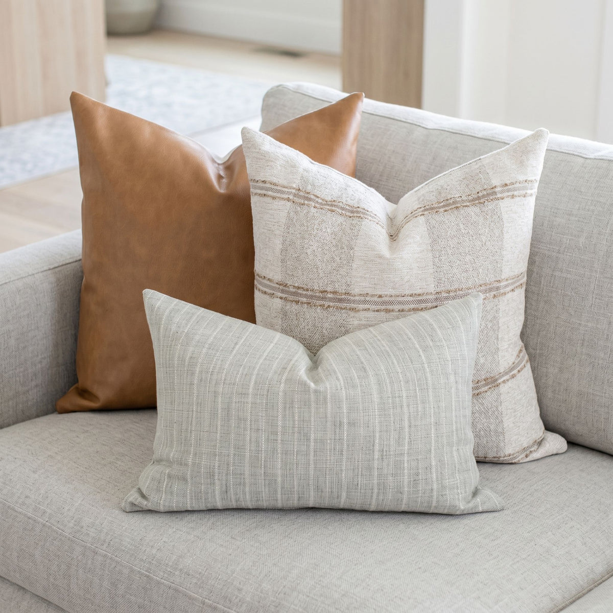 Set of three pillows including leather, rustic fabric, and gray striped lumbar on a neutral-toned sofa in a bright living space.